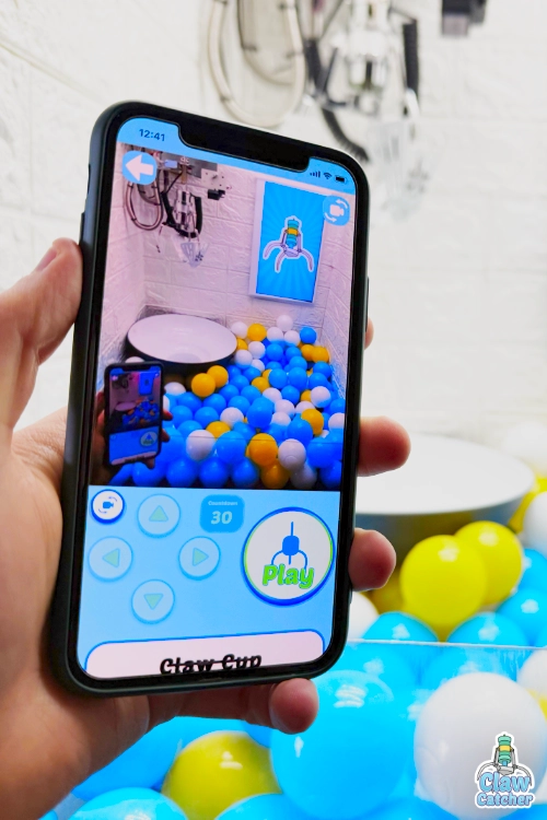 A person holding a smartphone playing the Claw Catcher web app, using on‑screen arrow controls to operate a real claw machine filled with colorful balls, with the physical claw machine visible blurred in the background.